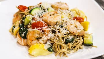 white plate with edamame linguini mixed with shrimps, zucchini, tomatoes, and shredded parmesan on top