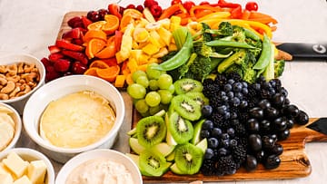 kiwi, apple, blueberries, strawberries, oranges, grapes, blackberries, peas, broccoli, cashews served on a wooden cutting board, with some cream in a white bowl