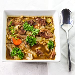 beef and barley soup served in a white bowl with a spoon on the side