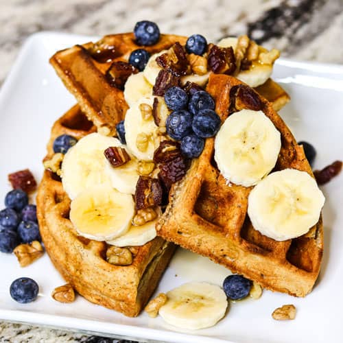 Spelt waffles with maple syrup, banana, blueberry, dates and walnuts on the top served on the white plate