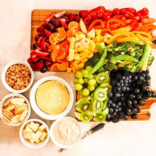 kiwi, apple, blueberries, strawberries, oranges, grapes, blackberries, peas, broccoli, served on a wooden cutting board, with some cashews and cream in a white bowl