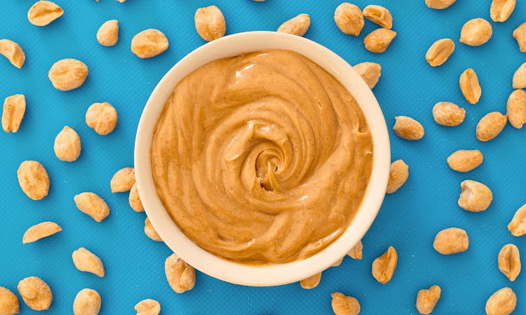 Peanut butter in a white bowl and some peanuts around