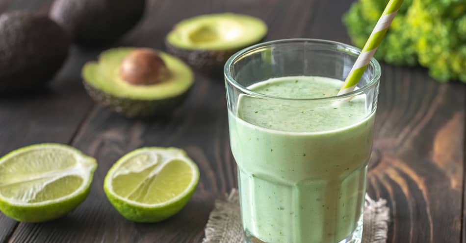 Glass full of green smoothie, avocado and limes on the side