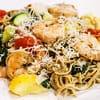white plate with edamame linguini mixed with shrimps, zucchini, tomatoes, and shredded parmesan on top