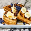 Spelt waffles with maple syrup, banana, blueberry, dates and walnuts on the top served on the white plate