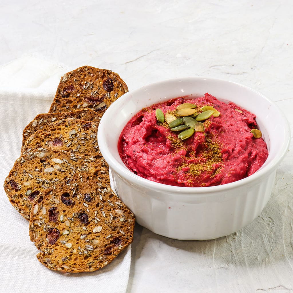 beet hummus served in a small white bowl, and some whole seed crackers on the side