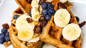 Spelt waffles with maple syrup, banana, blueberry, dates and walnuts on the top served on the white plate