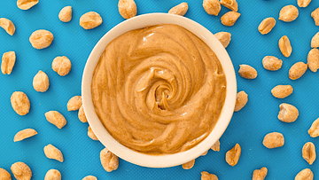 Peanut butter in a white bowl and some peanuts around