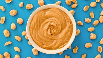 Peanut butter in a white bowl and some peanuts around