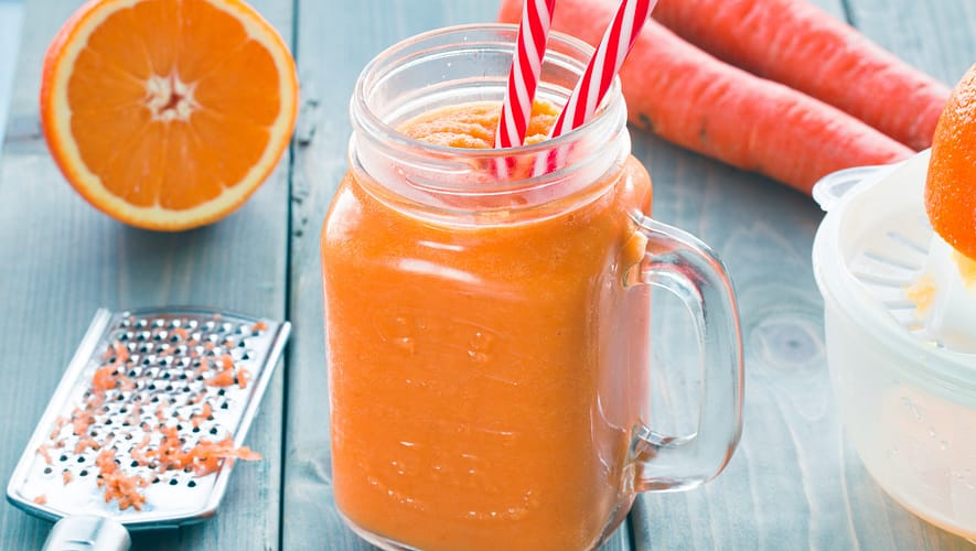 Orange smoothie served in a jar with two straws, and some carrots and oranges on the side